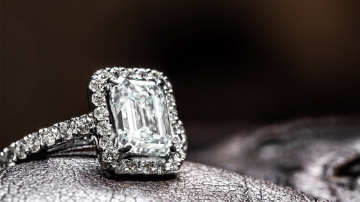 A luxurious emerald cut diamond engagement ring with a halo setting and diamond-encrusted band on a dark textured surface