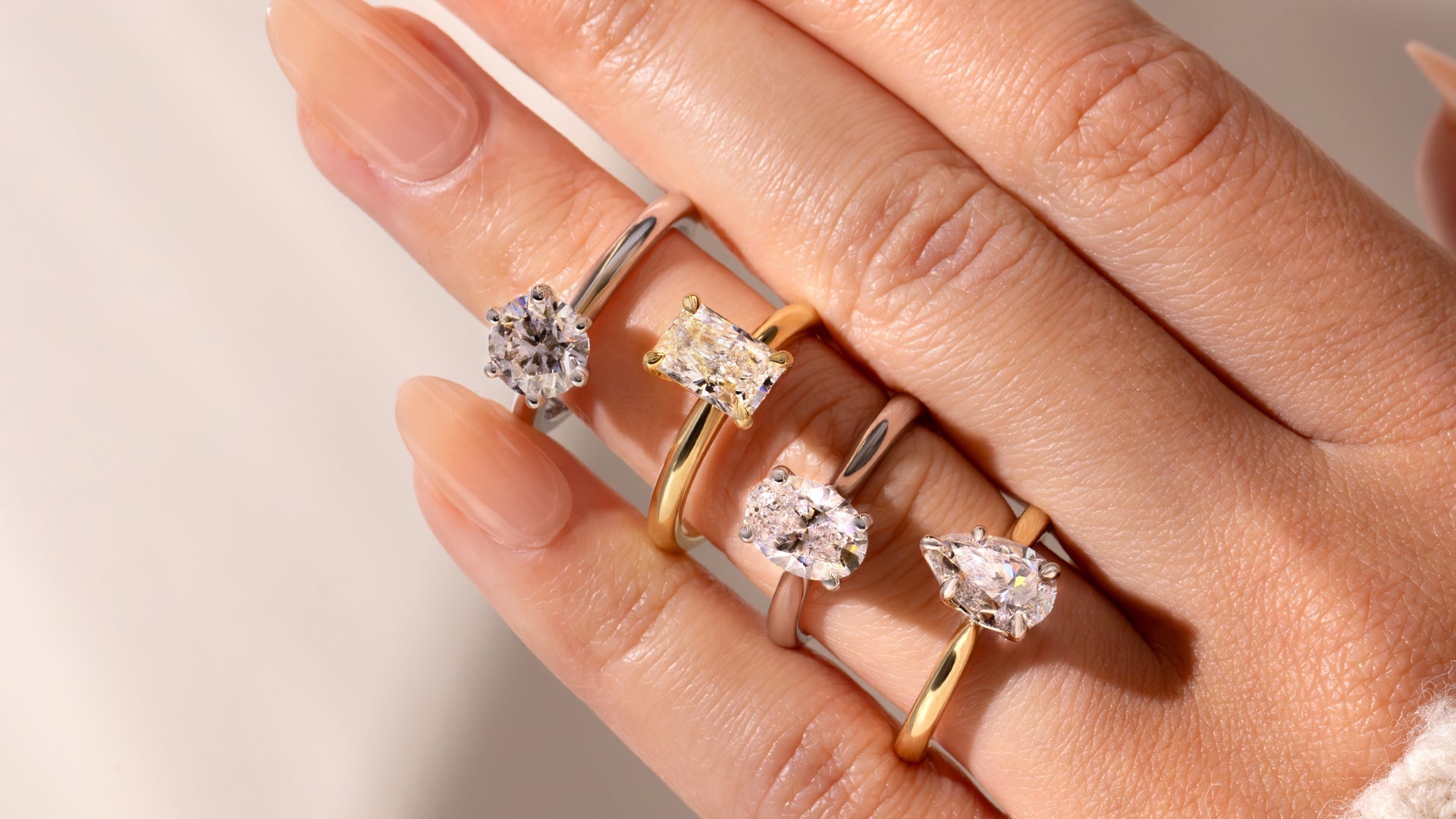 A hand adorned with four unique engagement rings, showcasing various diamond cuts set in gold and silver bands.