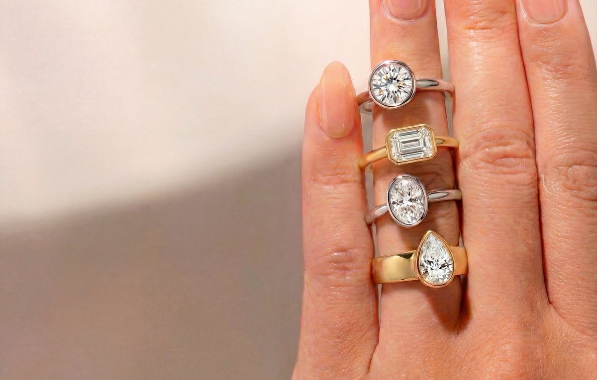 Hand wearing multiple bezel lab diamond engagement rings on a neutral background