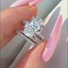 Oval-cut diamond bridal set featuring a four-prong engagement ring with a pavé band and a matching diamond wedding band, held against a white satin background.