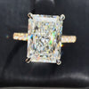 Radiant-cut diamond engagement ring featuring a four-prong setting and a sparkling pavé yellow gold band, shown against a dark textured background.