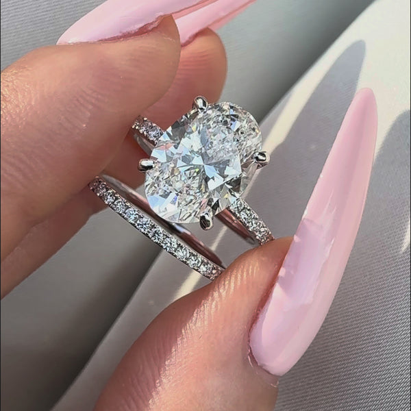 Oval-cut diamond bridal set featuring a four-prong engagement ring with a pavé band and a matching diamond wedding band, held against a white satin background.