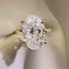 A large 3ct oval-cut lab diamond engagement ring set in a four-prong yellow gold band, resting on a cream-colored satin jewelry box liner.